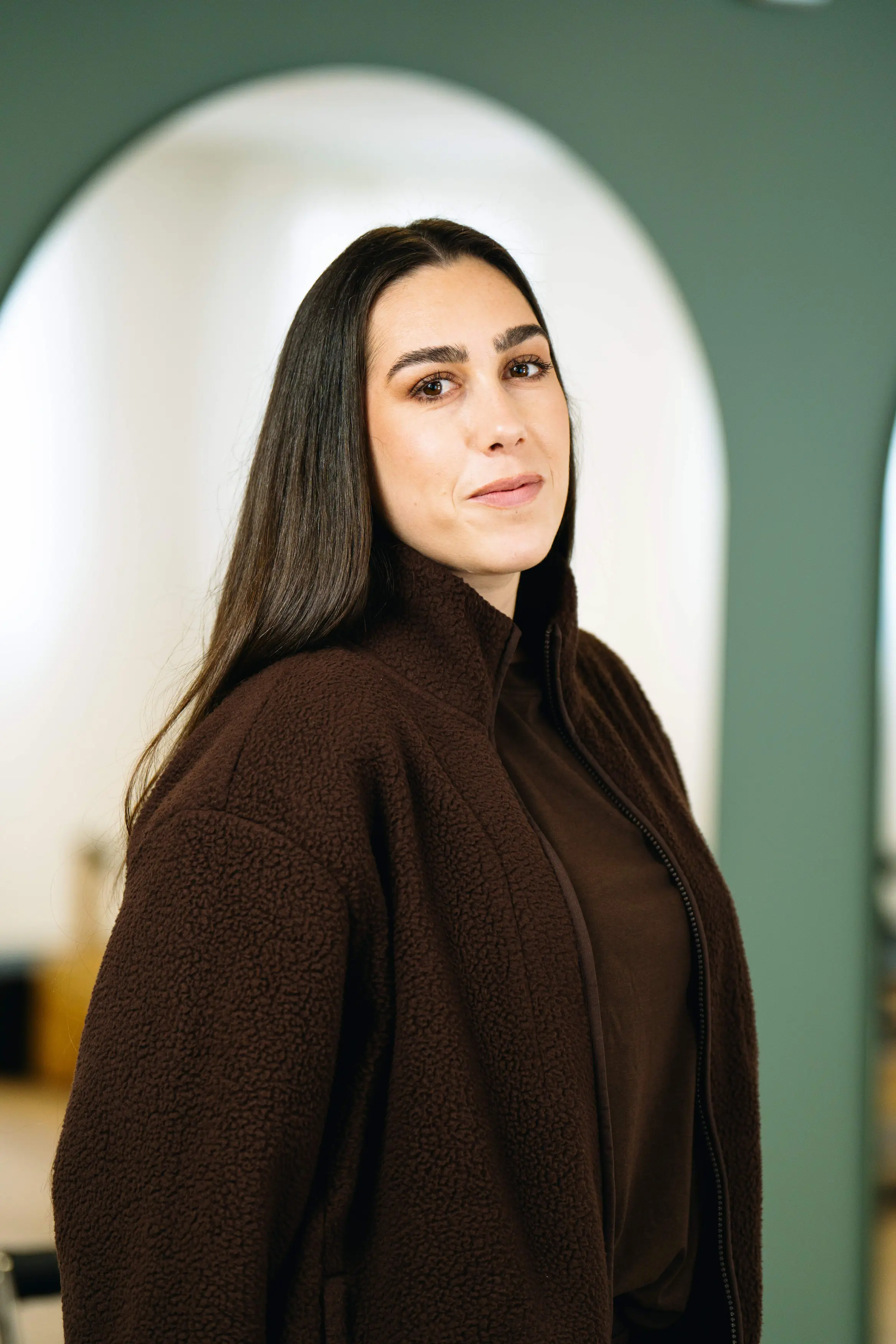 Portrait d'une femme souriante aux longs cheveux noirs portant une veste marron à col montant.