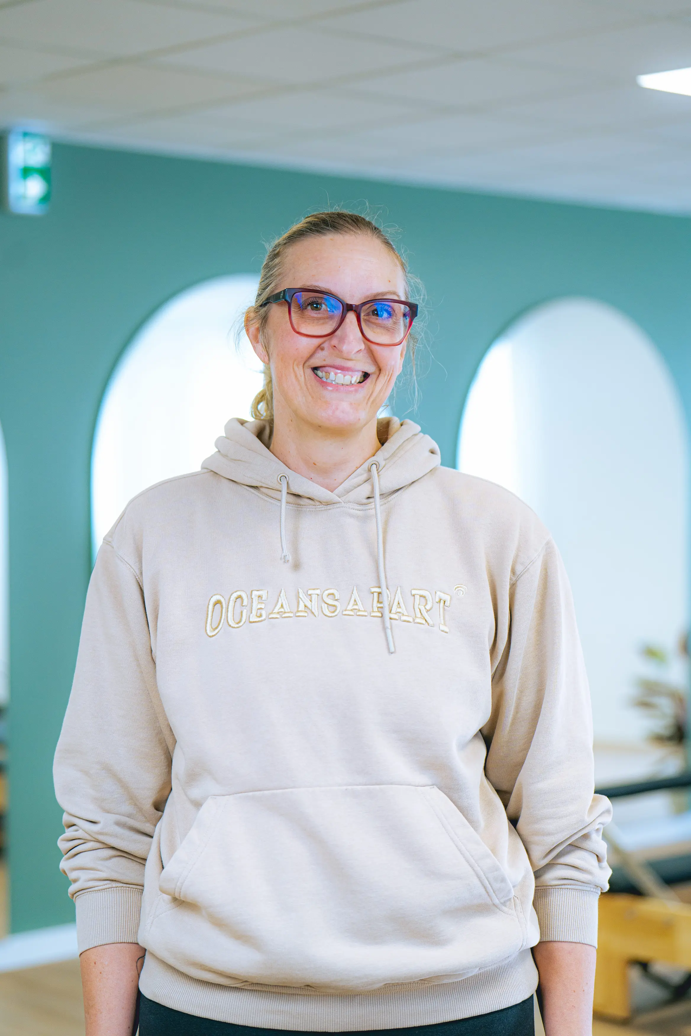 Femme souriante portant un sweat à capuche beige avec l'inscription « OCEANSAPART » et des lunettes rouges dans un intérieur avec des murs verts.