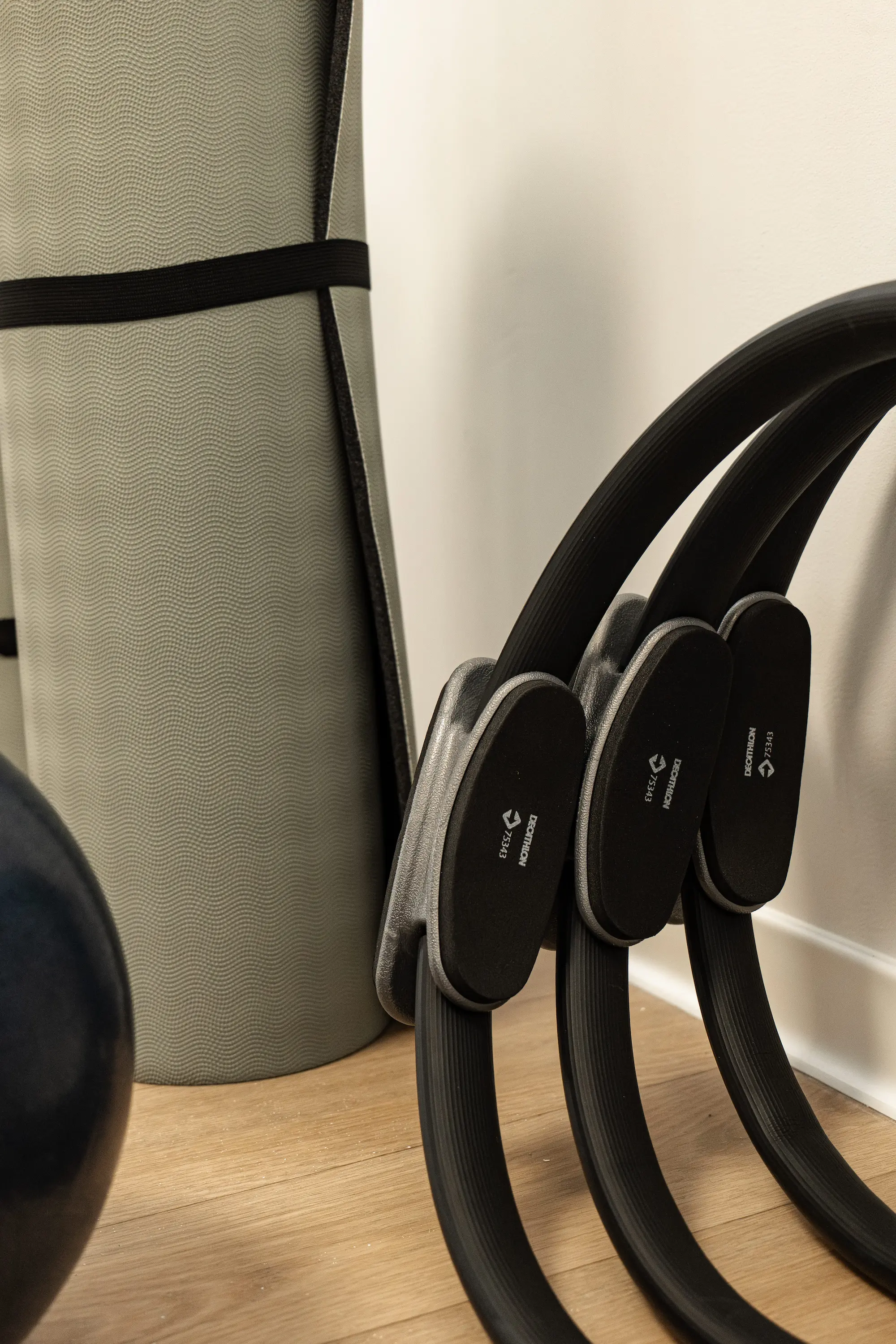 Trois cercles de Pilates noirs appuyés contre un mur avec un tapis de yoga gris roulé à côté.