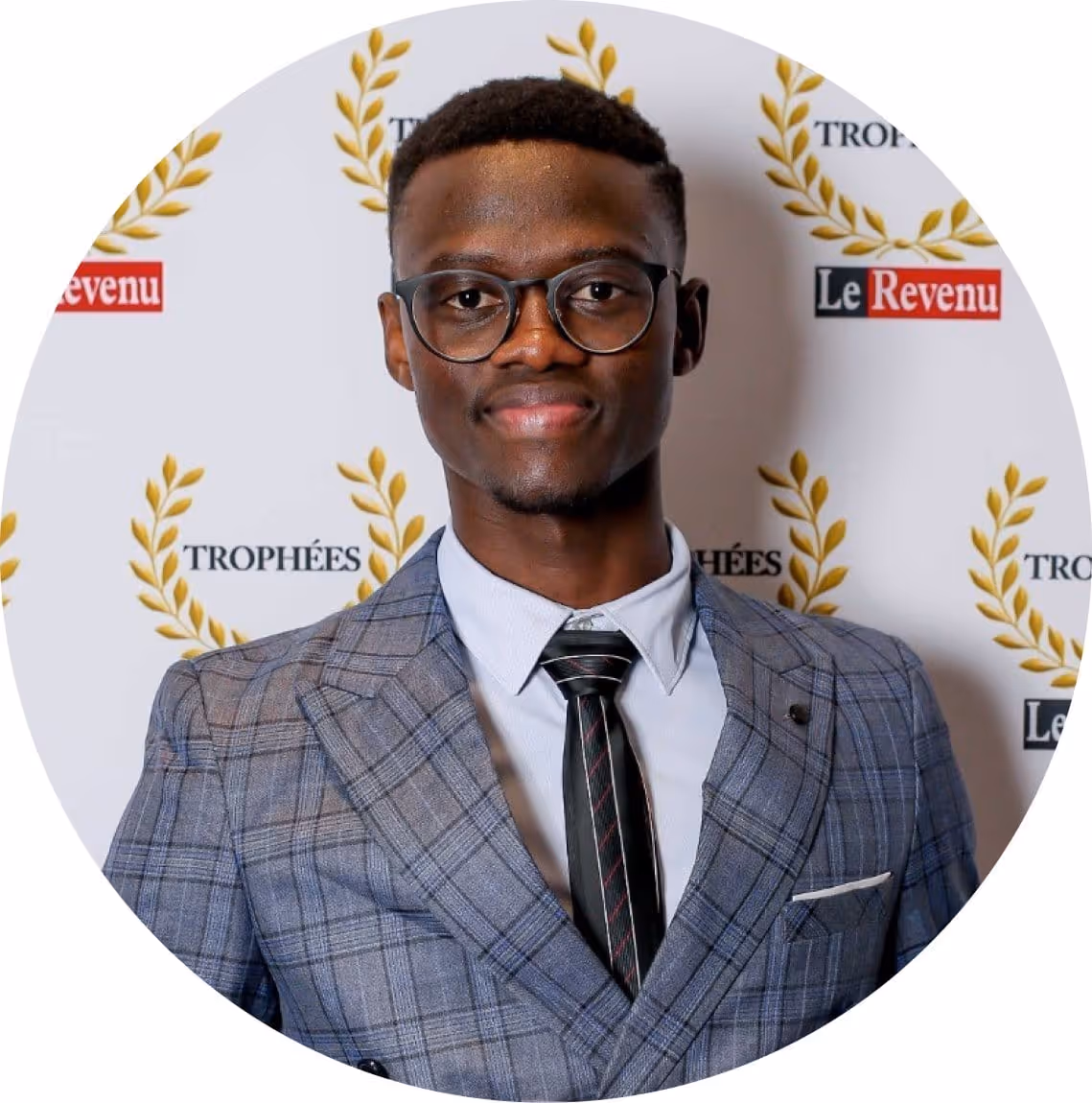Jeune homme portant un costume à carreaux gris, chemise blanche, cravate noire et lunettes, souriant.