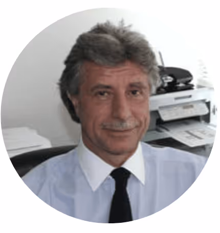 Homme d'âge moyen souriant, portant une chemise blanche et une cravate noire, assis dans un bureau.