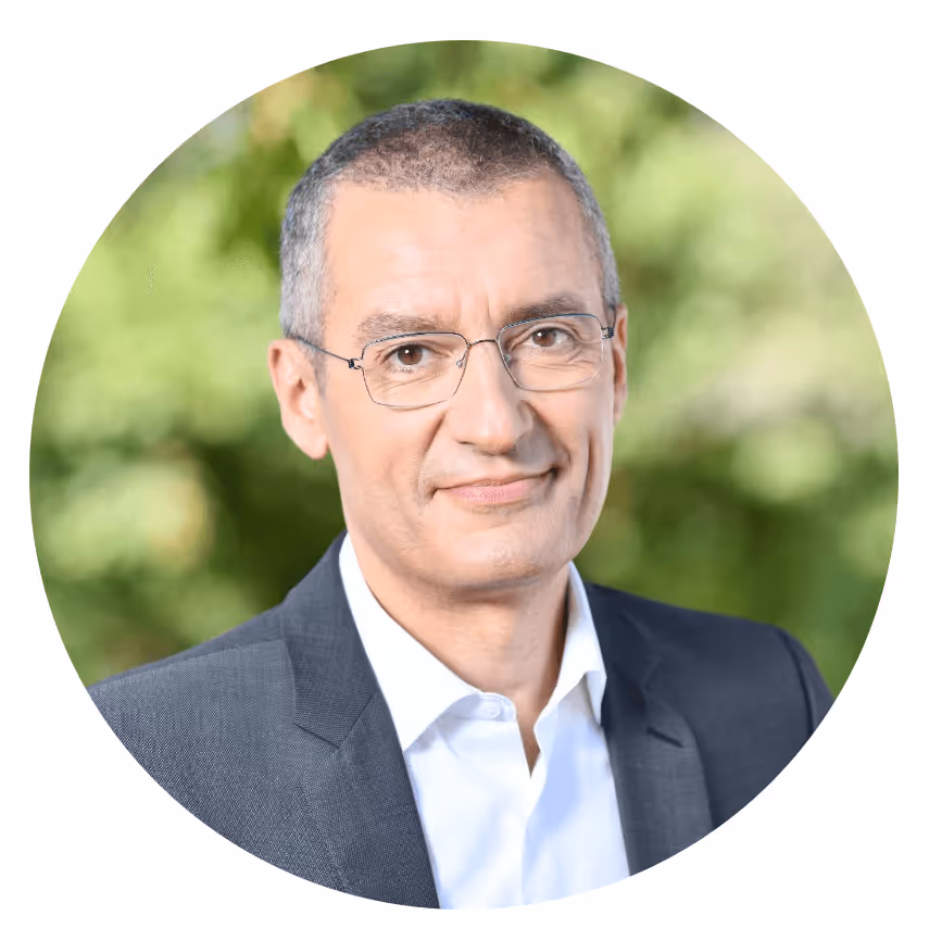 Homme portant un costume bleu et des lunettes souriant devant un fond flou de verdure.