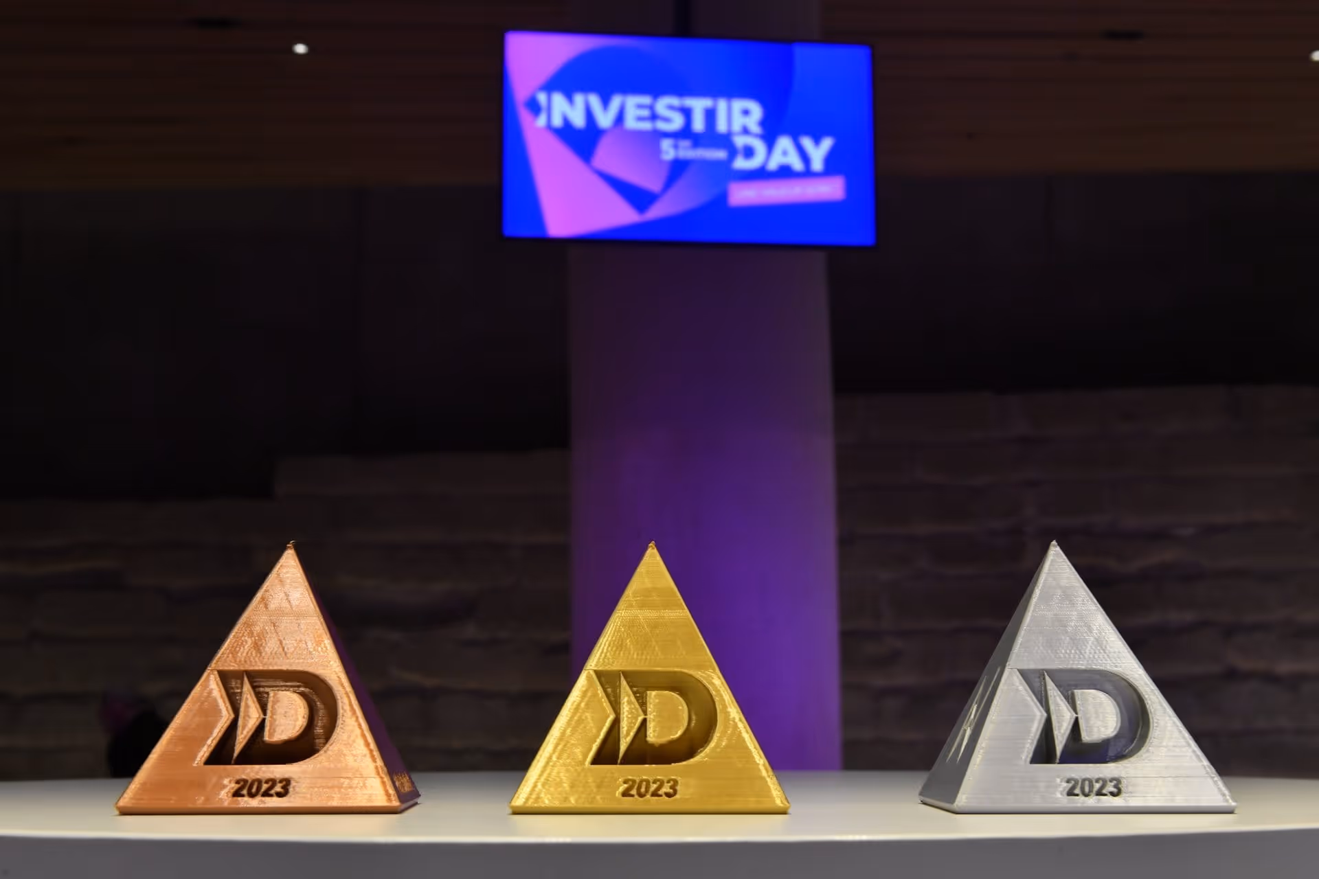 Trois trophées pyramides 2023 en bronze, or et argent alignés sur une table sous un écran LED.