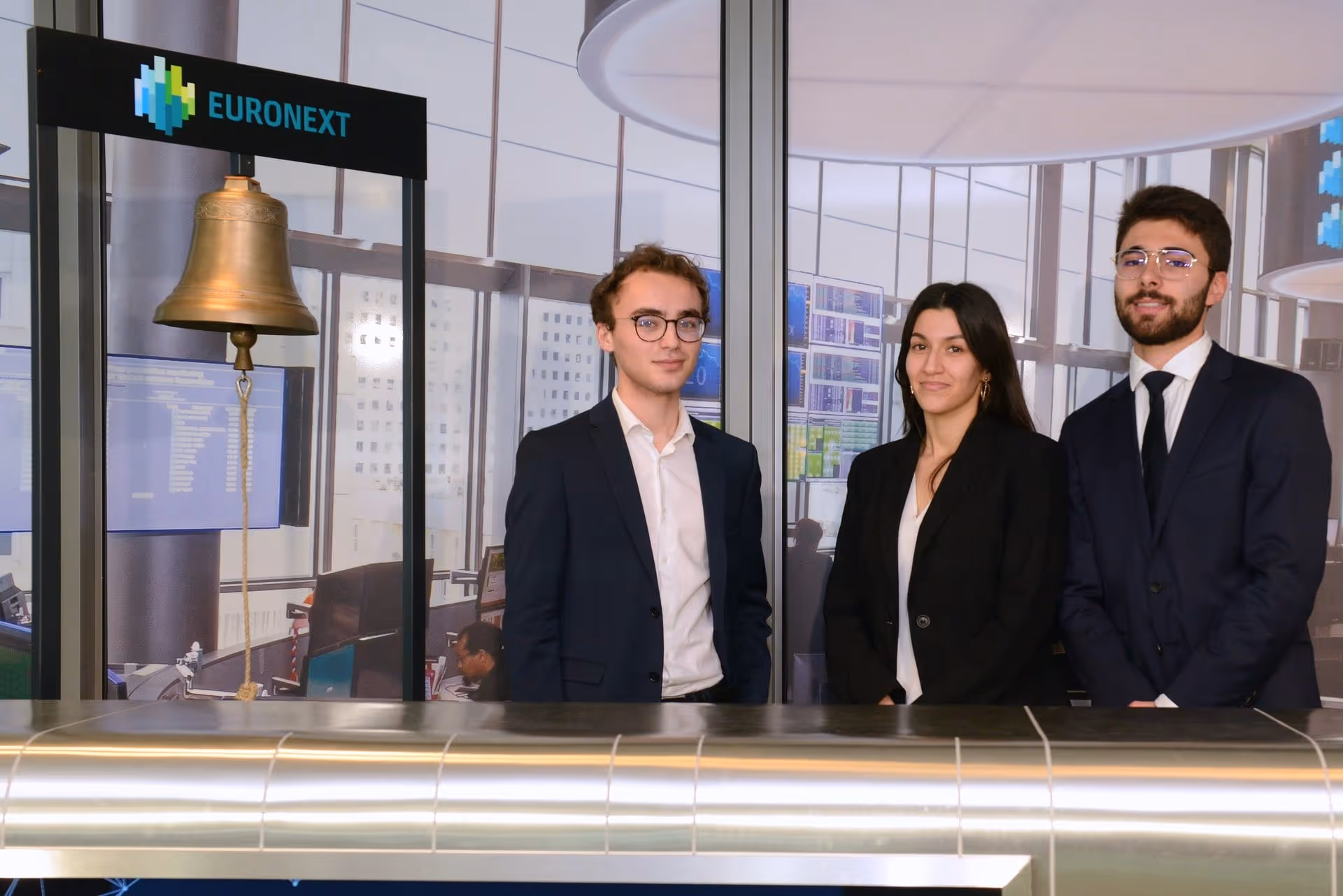 Trois jeunes professionnels en costume debout devant une cloche Euronext dans un bureau moderne.