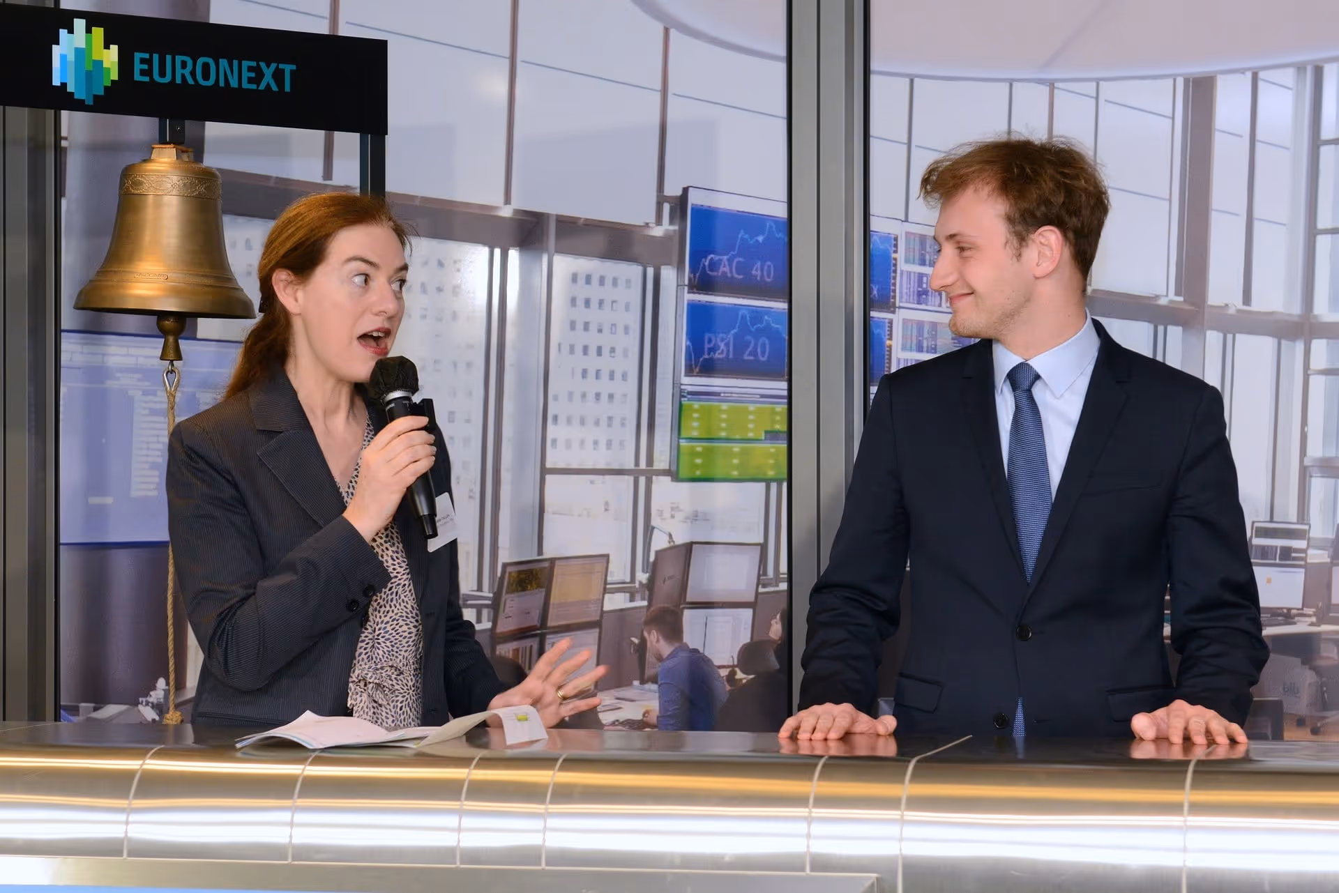 Deux personnes en costume discutant près d'une cloche avec un panneau Euronext en arrière-plan.
