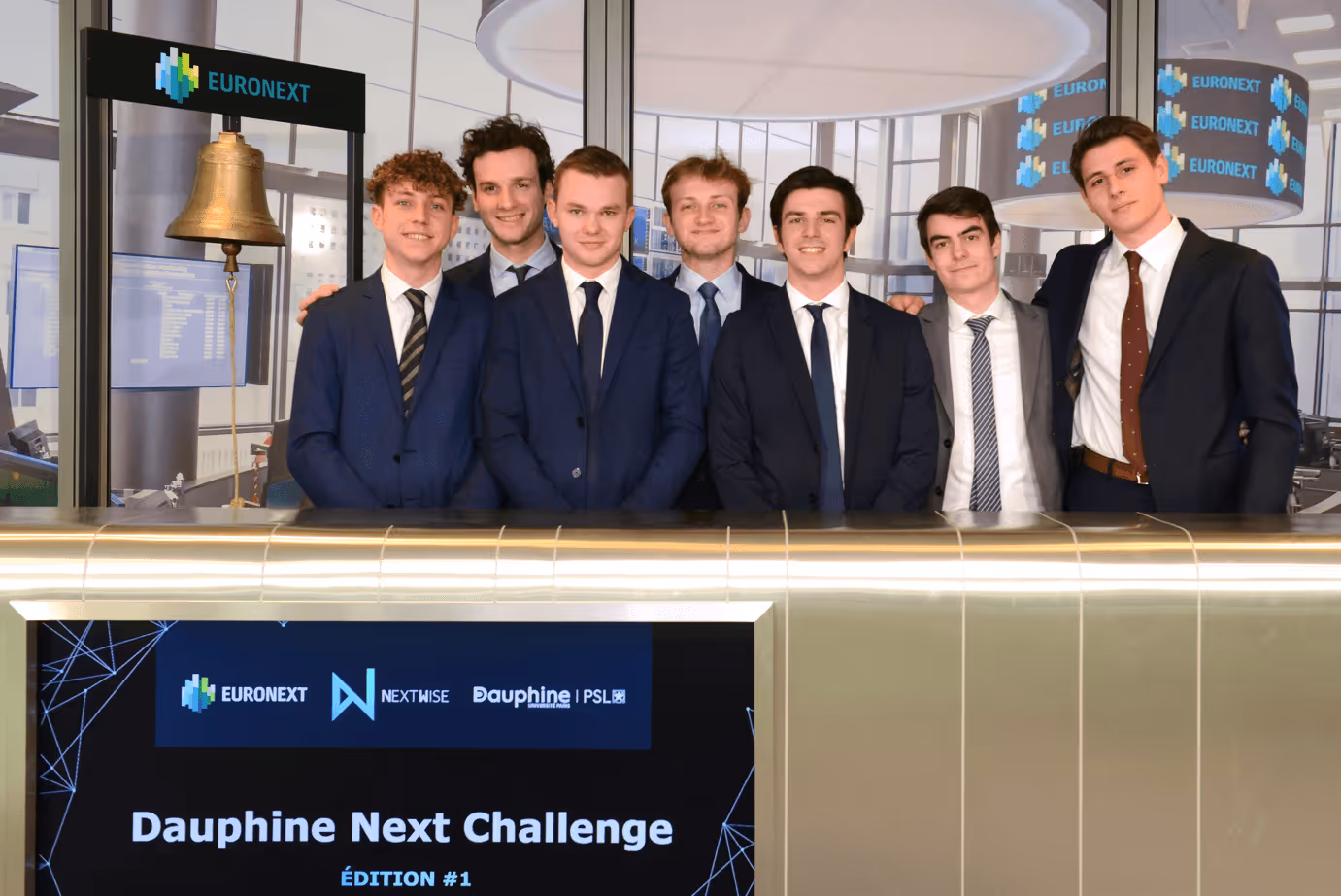 Sept jeunes hommes en costume posent derrière un comptoir avec un écran et une cloche Euronext en arrière-plan.