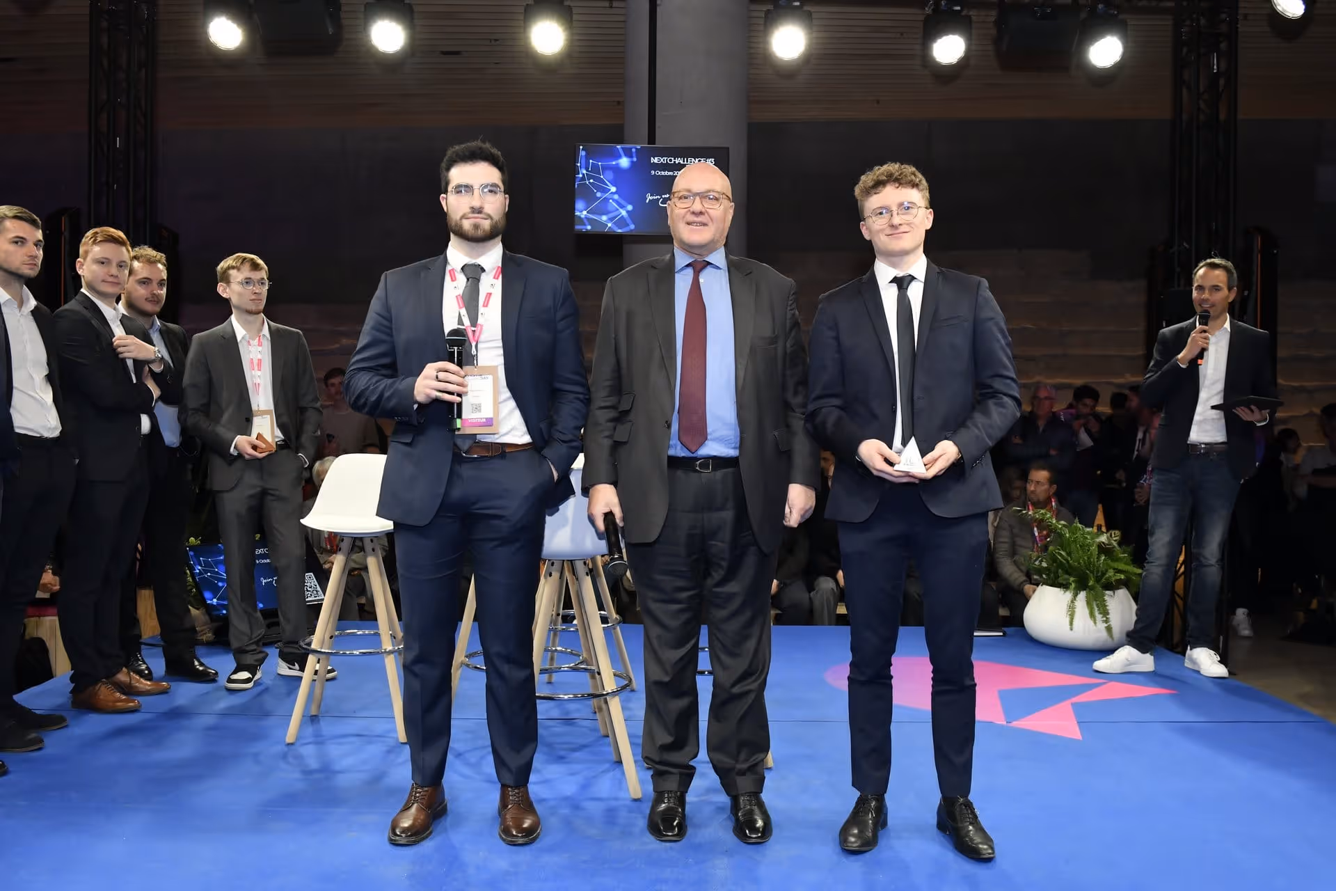Trois hommes en costume debout sur scène avec microphones et trophée lors d'un événement professionnel.