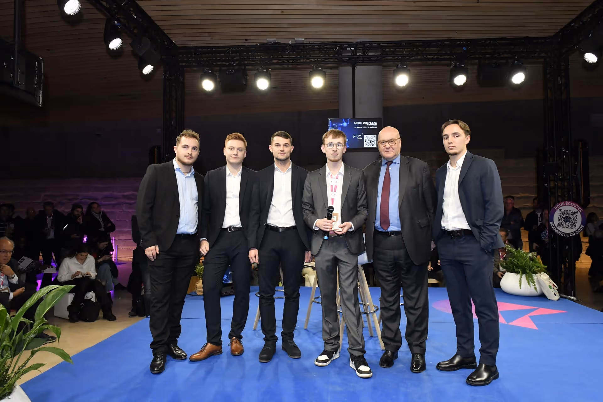 Six hommes en costume debout sur une scène bleue lors d'un événement formel.