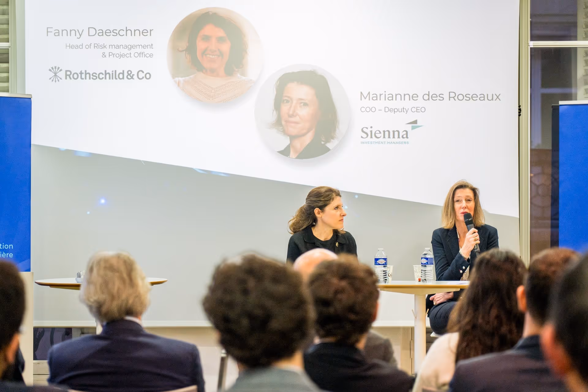 Deux femmes assises derrière une table parlent devant un public, avec des noms et photos projetés au mur.