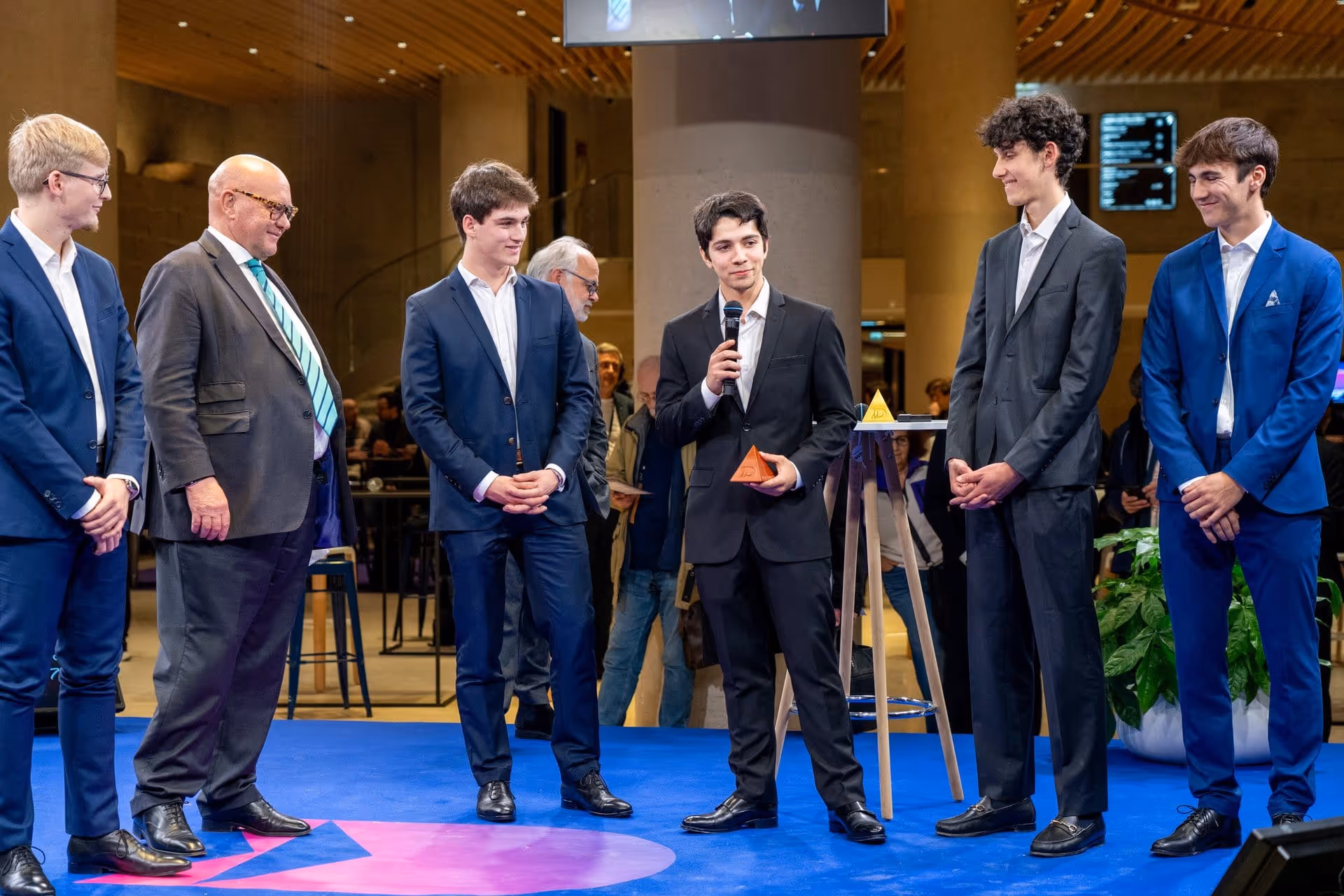 Un groupe d'hommes en costume sur une scène, un homme tenant un micro et un trophée triangulaire.