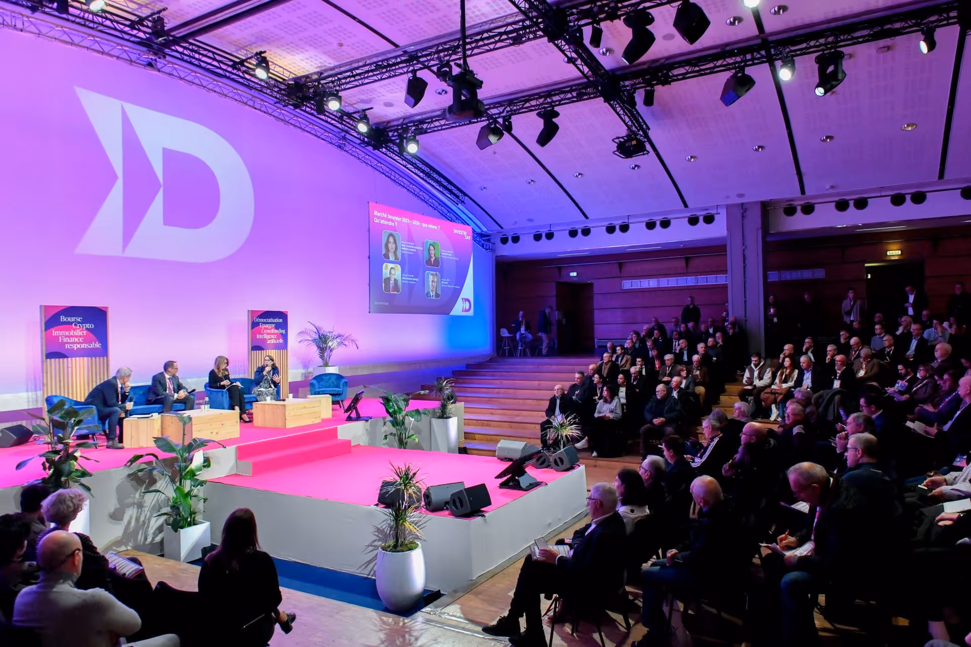 Conférence avec quatre intervenants sur scène et un large public dans une salle éclairée en violet.