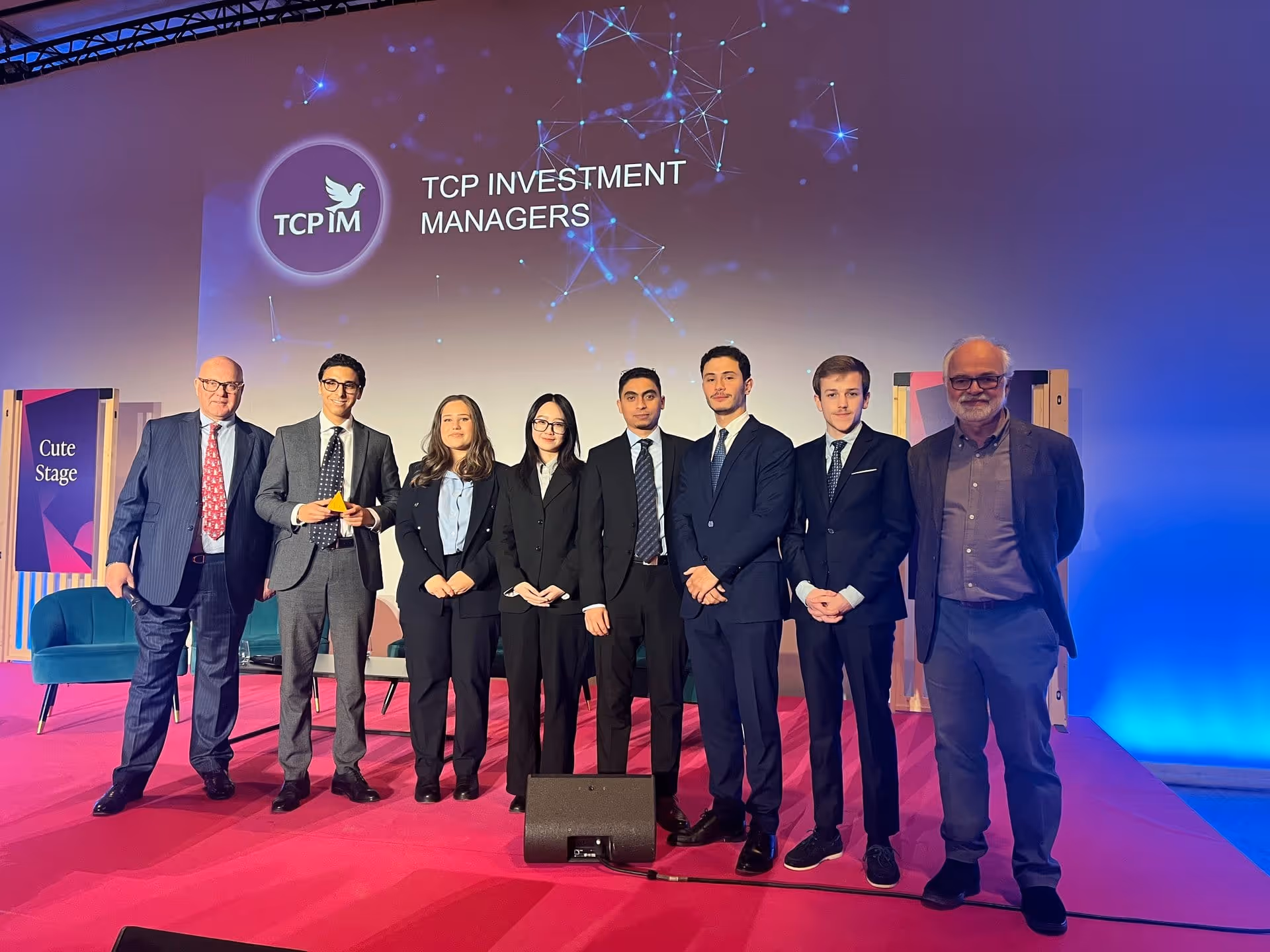 Groupe de sept personnes en costume sur une scène rose avec le logo TCP Investment Managers en arrière-plan.