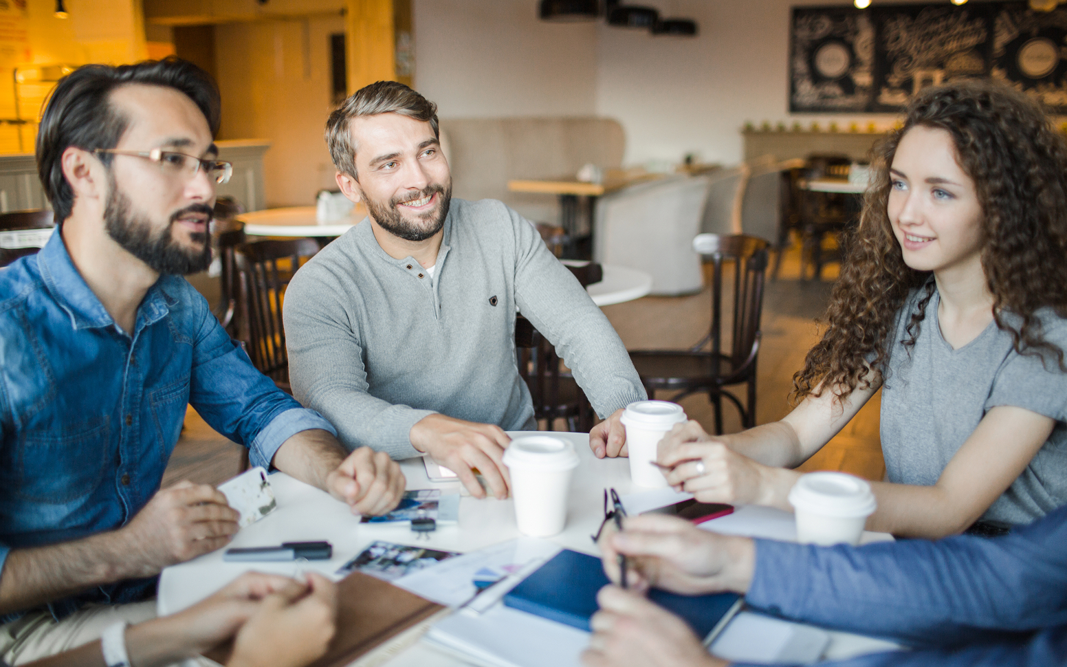 Group of young adults sitting around a table in a cafe, smiling and talking with coffee cups and documents in front of them.