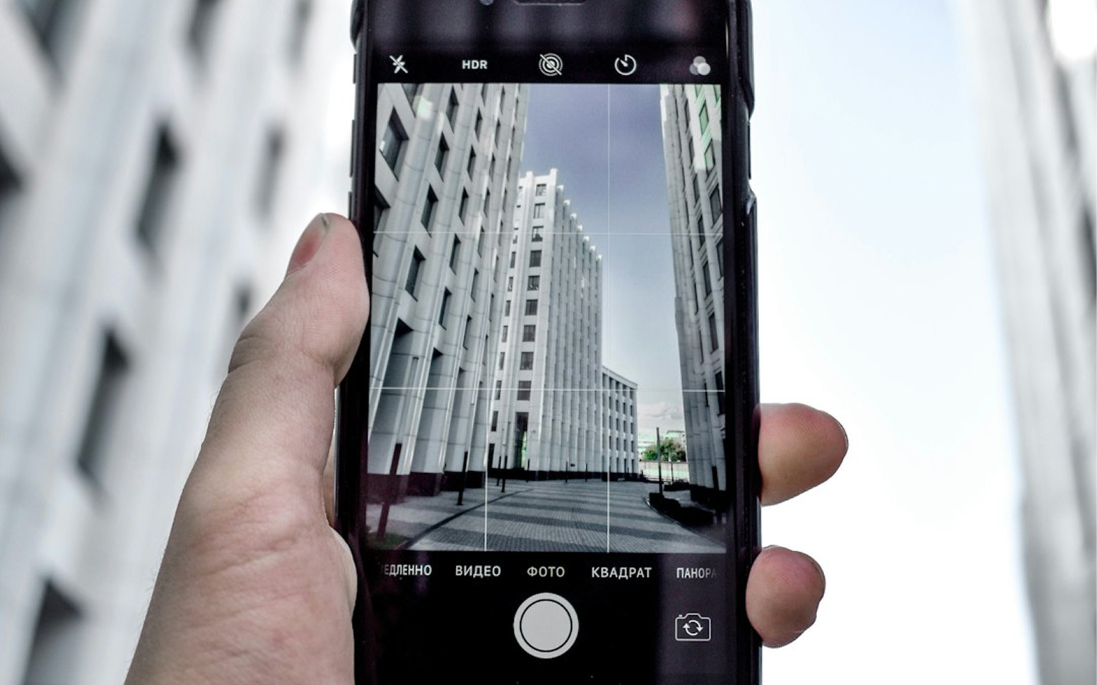 Hand holding a smartphone capturing photo of modern tall white buildings between a paved walkway under a clear sky.