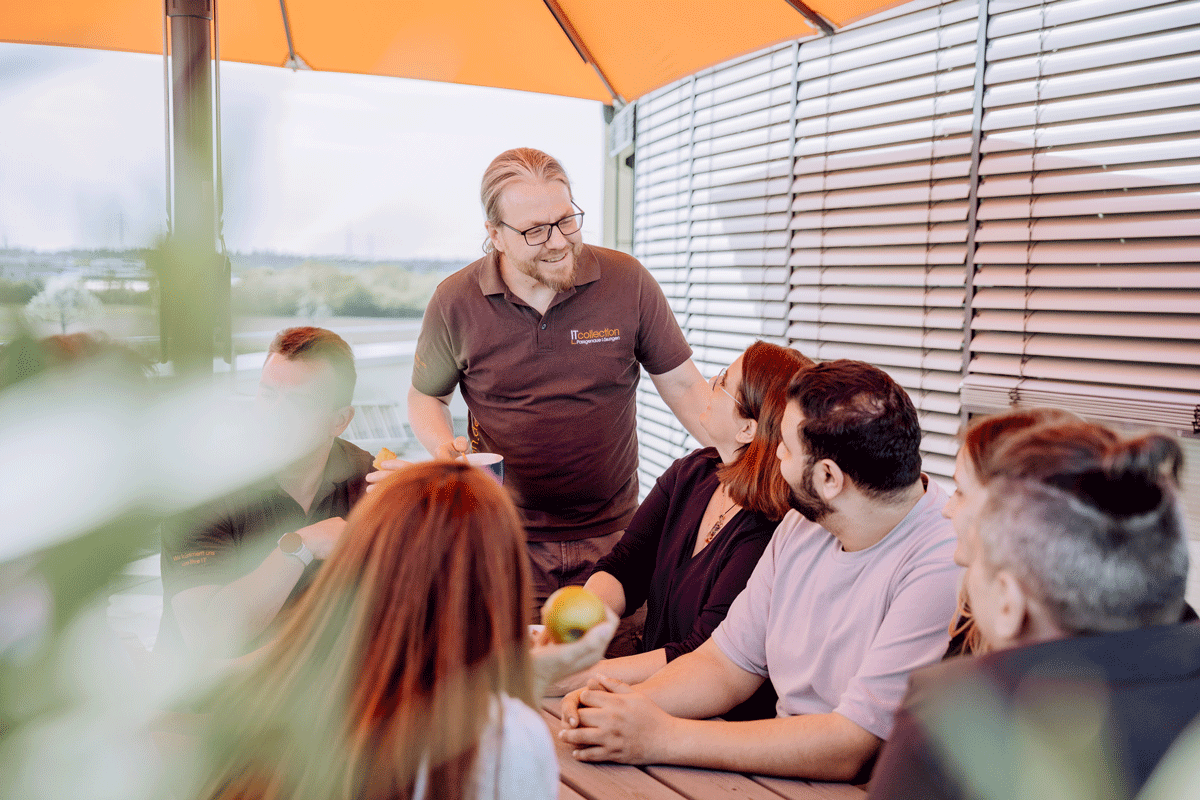 Gruppe von sechs Personen bei einem zwanglosen Treffen im Freien an einem Tisch unter einem orangefarbenen Sonnenschirm.