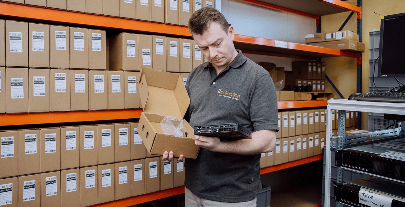 Mann in grauem Hemd begutachtet ein elektronisches Gerät in einem Lagerhaus mit beschrifteten Kartons in orangefarbenen Regalen.