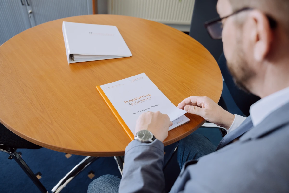Ein Mann sitzt an einem runden Holztisch und liest in einer Broschüre mit einem Projektvertrag, die den Titel „ITco Full Service“ trägt.