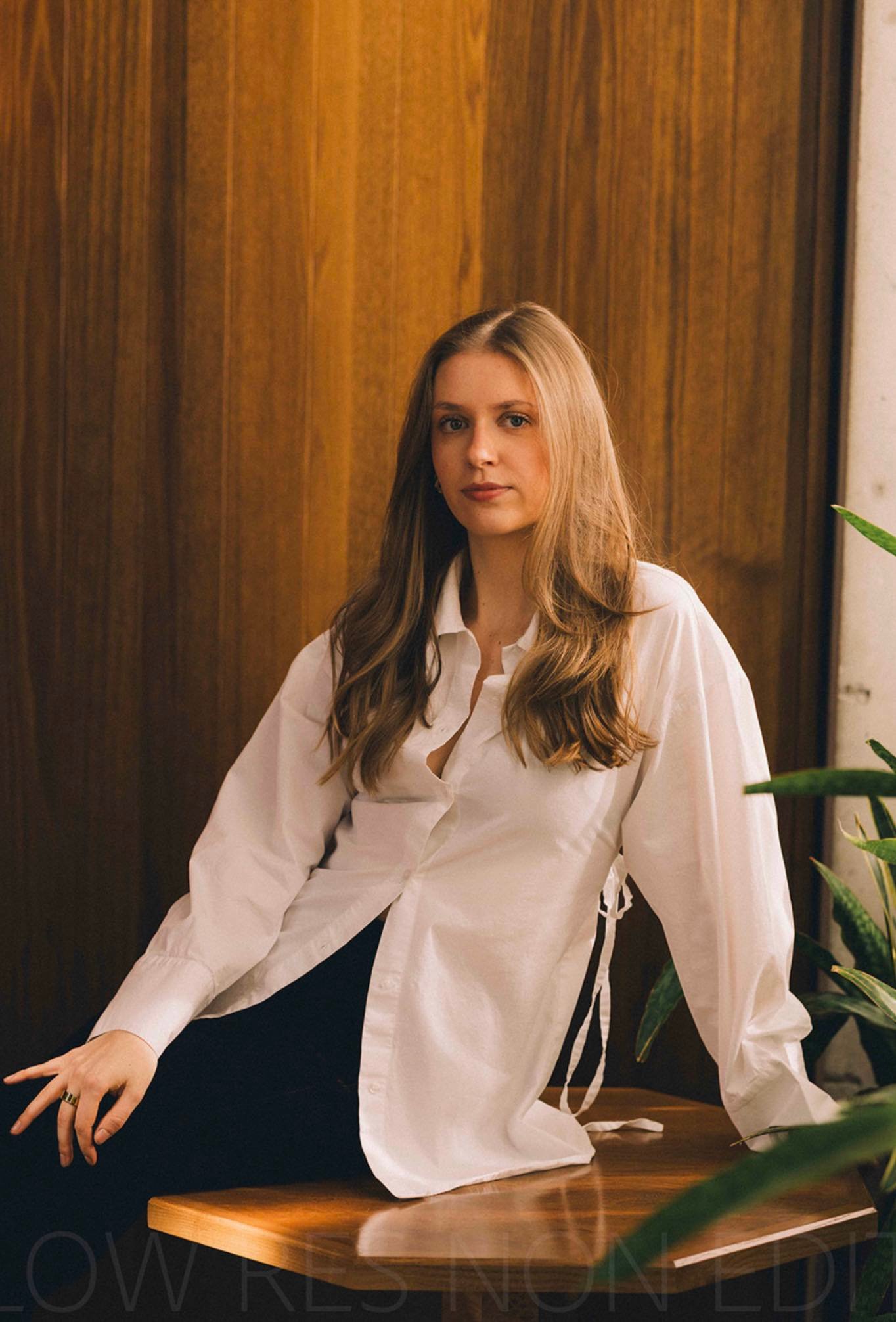 Jonge vrouw met los hangend lang haar, zittend op een houten tafel voor een houten achtergrond, gekleed in een witte blouse en zwarte broek.