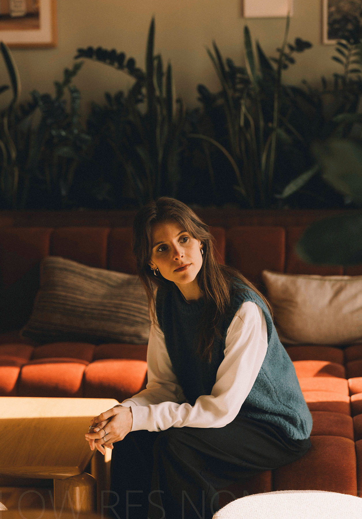Vrouw die op een oranje bank zit met planten op de achtergrond, kijkend naar rechts.