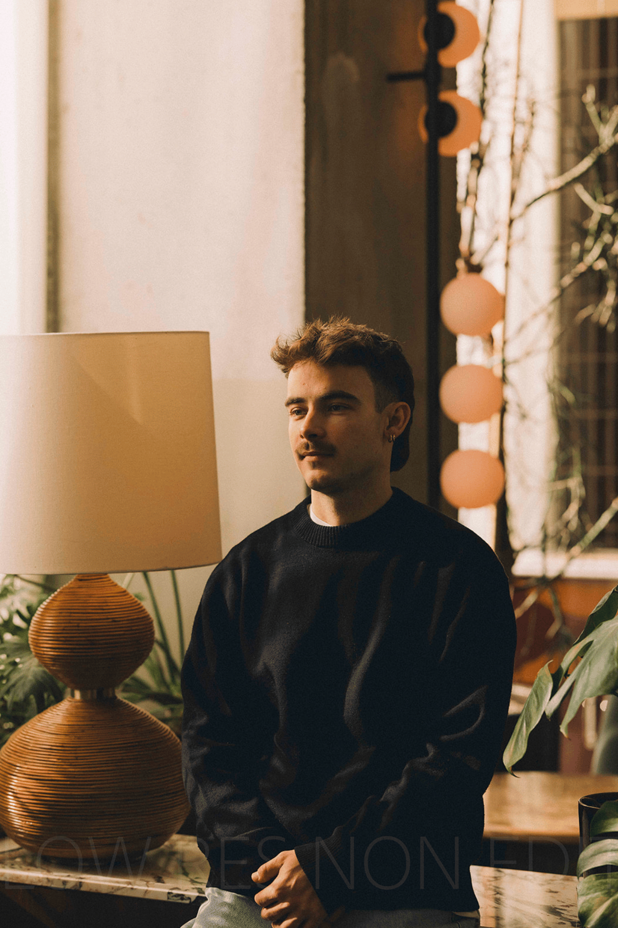 Man met een zwarte trui zittend bij een tafellamp en kamerplanten in een warme, sfeervolle kamer.