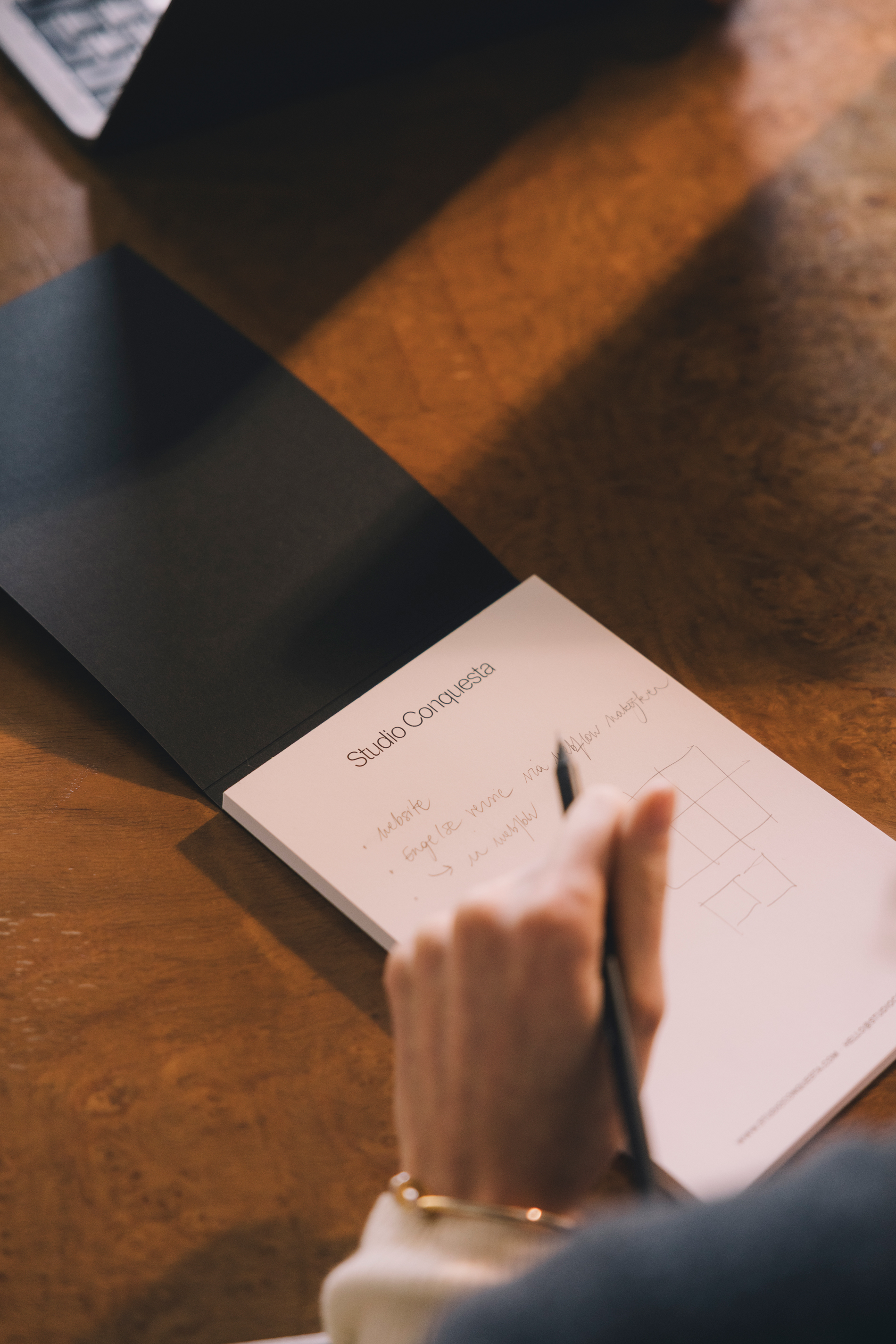 Iemand schrijft aantekeningen op een notitieblok met de koptekst Studio Conquesta op een houten tafel.