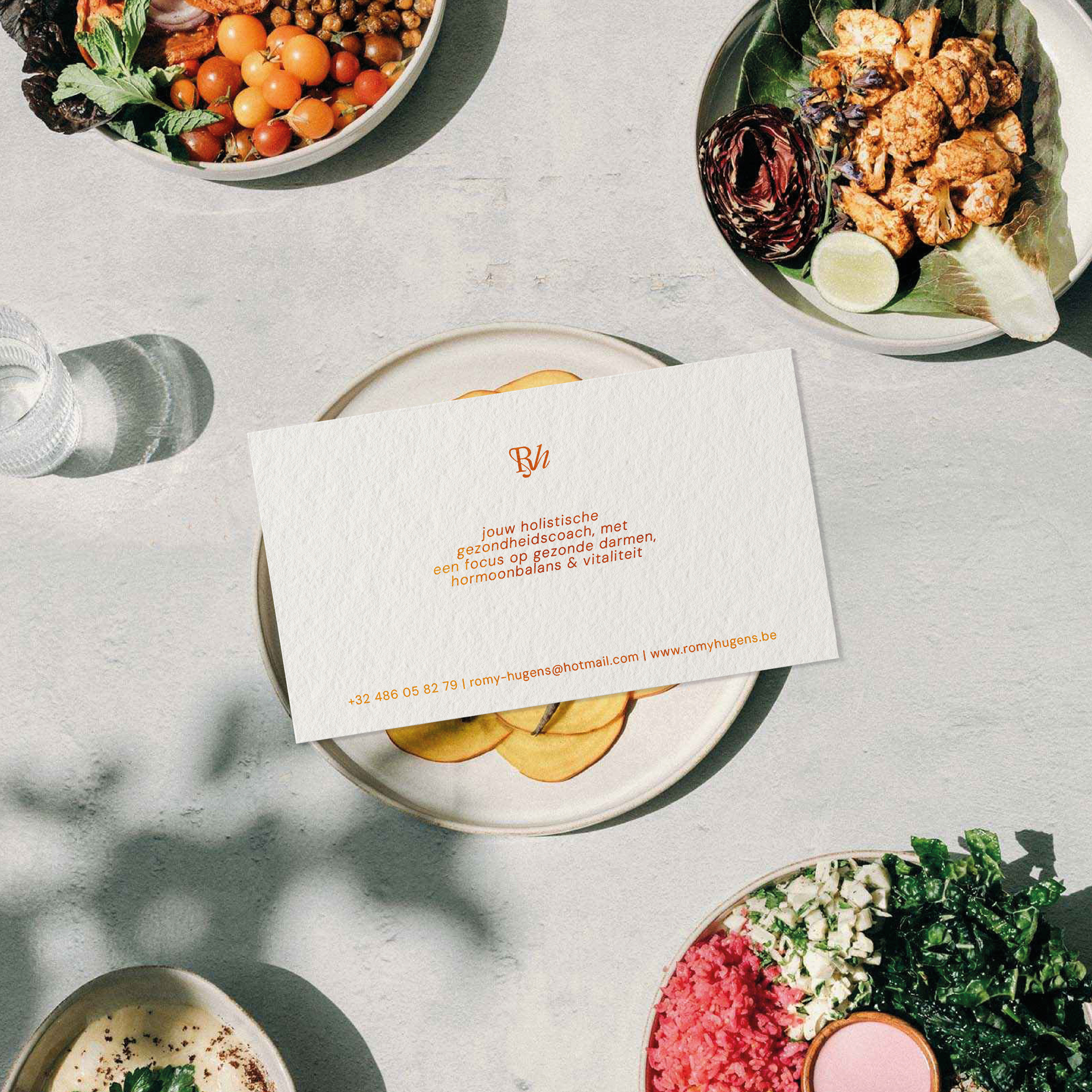 Visitekaartje met tekst over een holistische gezondheidscoach ligt op een bord met appels, omgeven door vier verschillende borden met kleurrijke gezonde maaltijden.