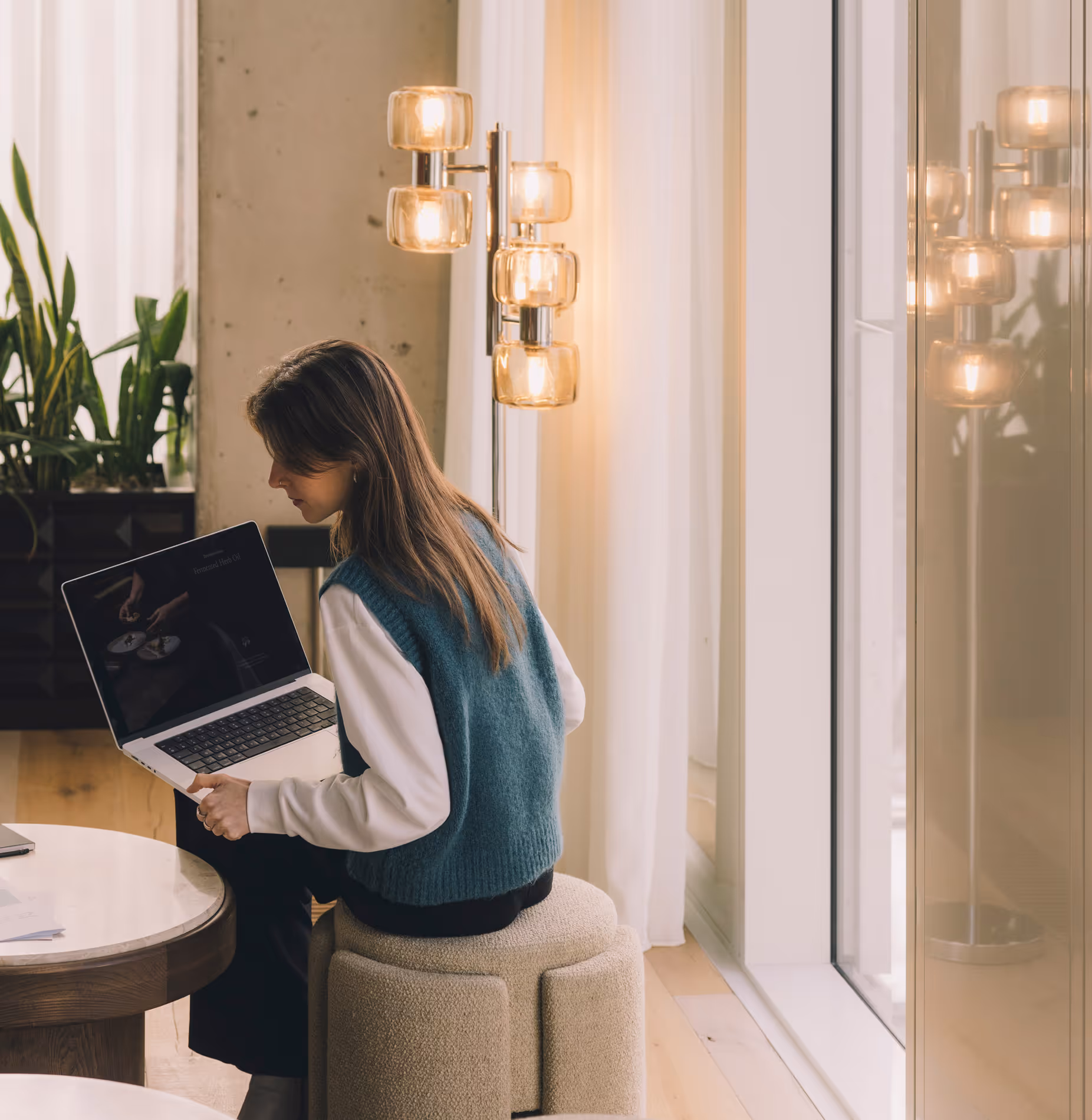 Vrouw zittend op een krukje in een lichte kamer, werkend op een laptop.