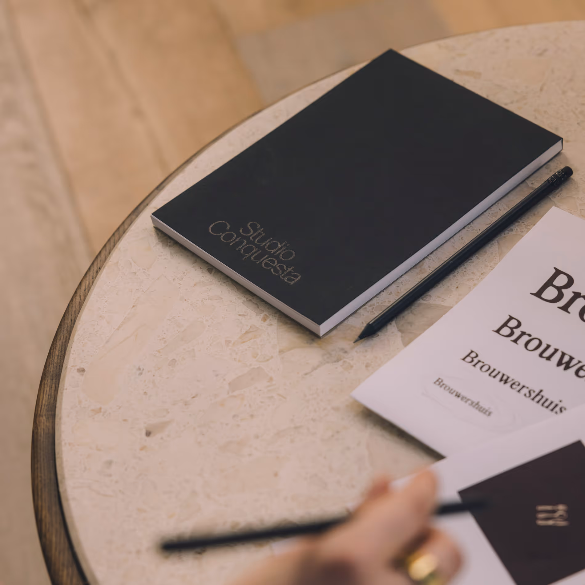 Een zwarte notitieboekje met tekst 'Studio Conquesta' op een ronde tafel met een potlood en een stuk papier.