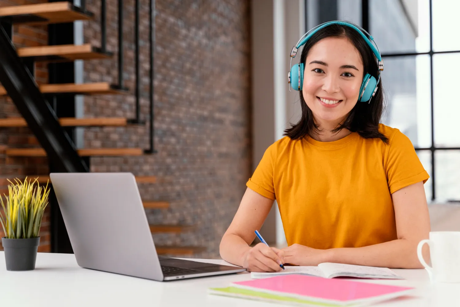 Young woman in orange shirt with blue headphones smiling while studying with notebook and laptop at a white desk.