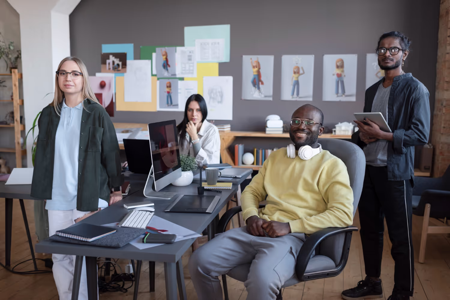 Four diverse young professionals in a modern office, two standing and two seated near desks with computers and office supplies.