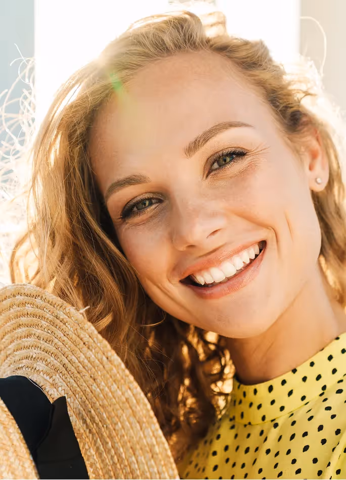Close-up of a smiling woman with curly blonde hair holding a straw hat, wearing a yellow polka dot top, with sunlit background.