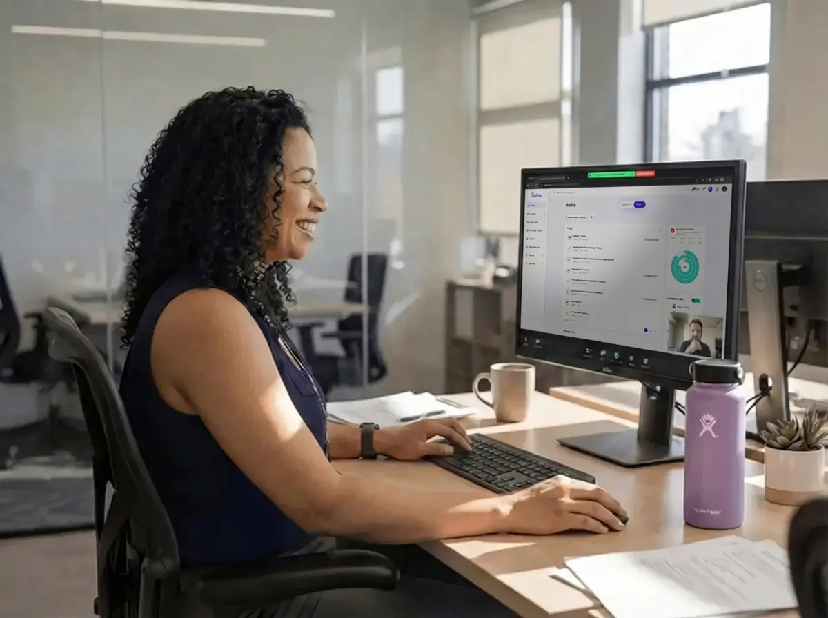 Smiling woman with curly hair working on a desktop computer in a modern office setting, with a purple water bottle and coffee mug on the desk.
