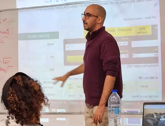 Man in glasses and maroon sweater pointing at a projection screen while explaining to a seated person in a classroom setting.