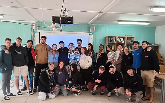 Group of twenty young adults posing together in a classroom with a projector and bookshelves in the background.