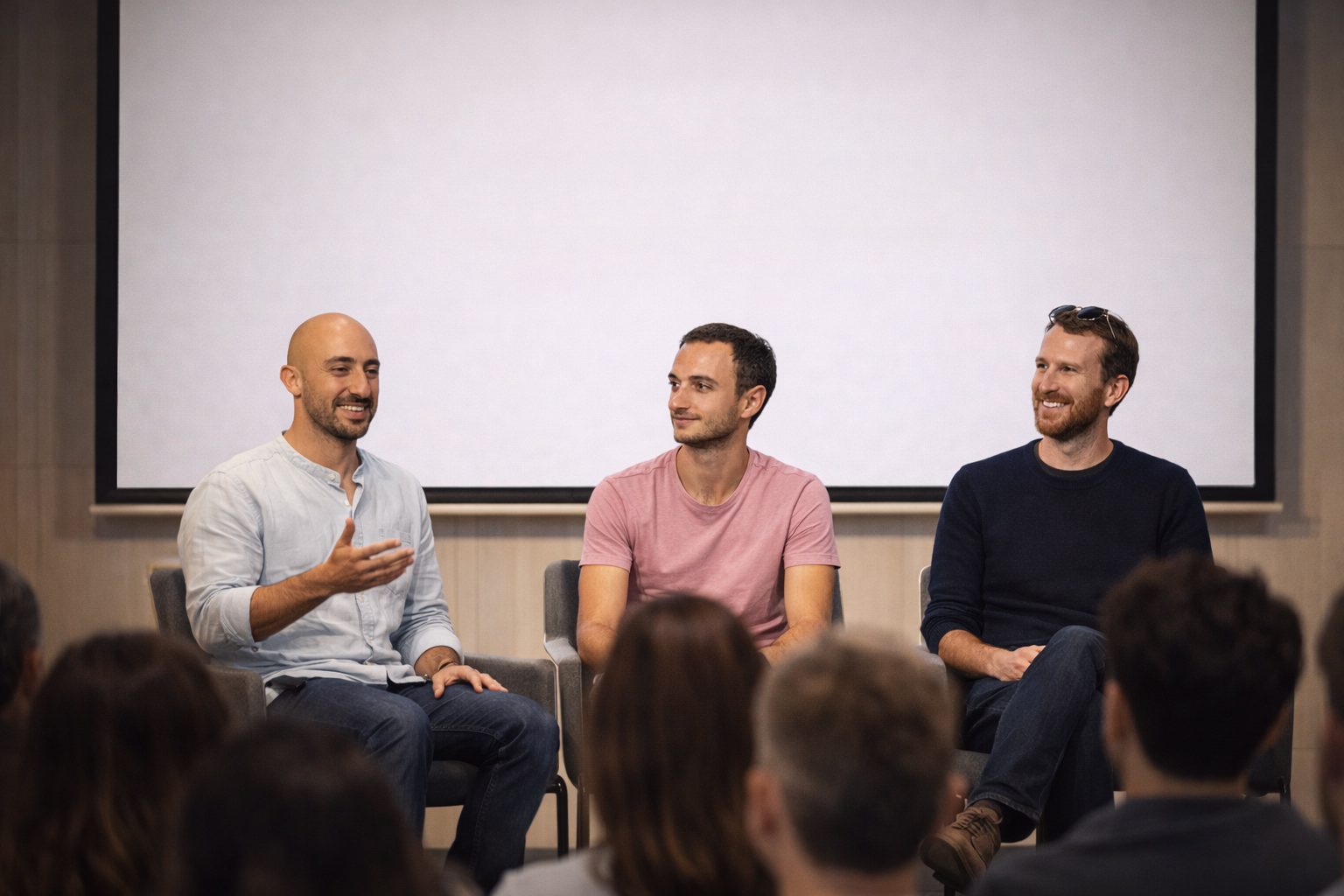Three men on a panel stage in front of a projection screen, speaking to an audience at an event.