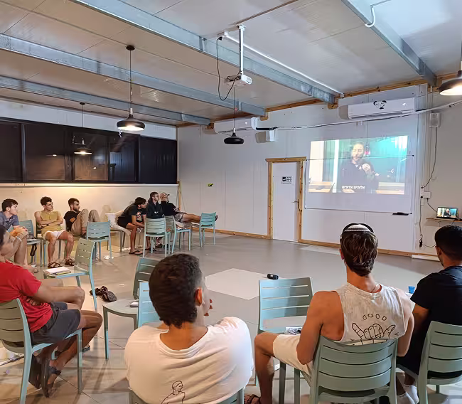 Group of people sitting on chairs in a room watching a man speaking on a projected video screen.