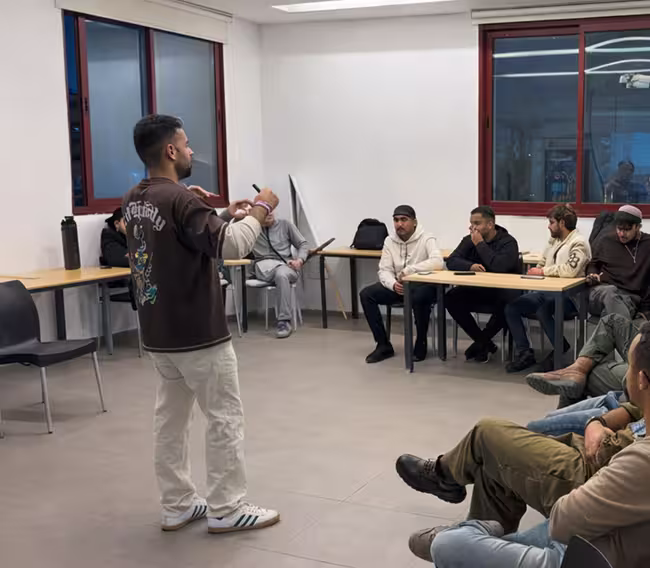 A man standing and speaking to a group of seated men in a classroom or meeting room.