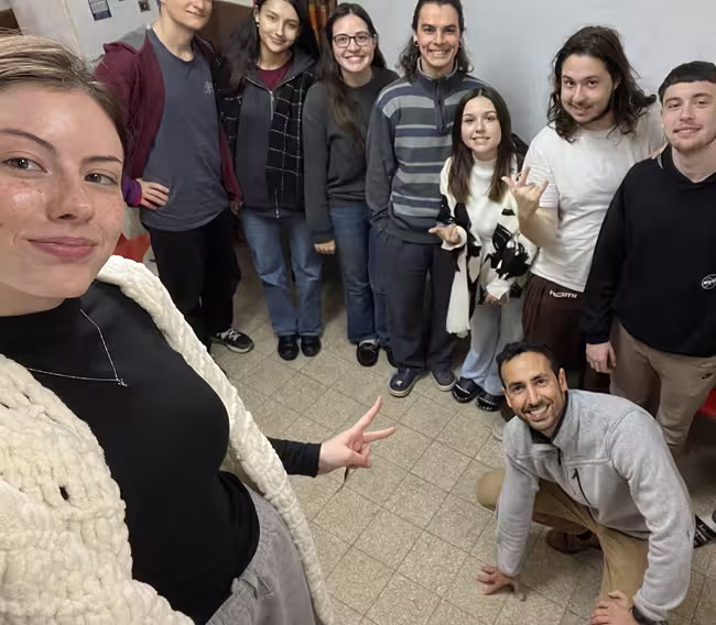 Group selfie of nine young adults smiling indoors, with one person crouching on the floor and others standing close together.
