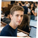 Young man with short brown hair sitting at a desk with an open laptop in a classroom.