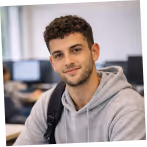 Young man with curly hair wearing a gray hoodie and backpack in an office environment.