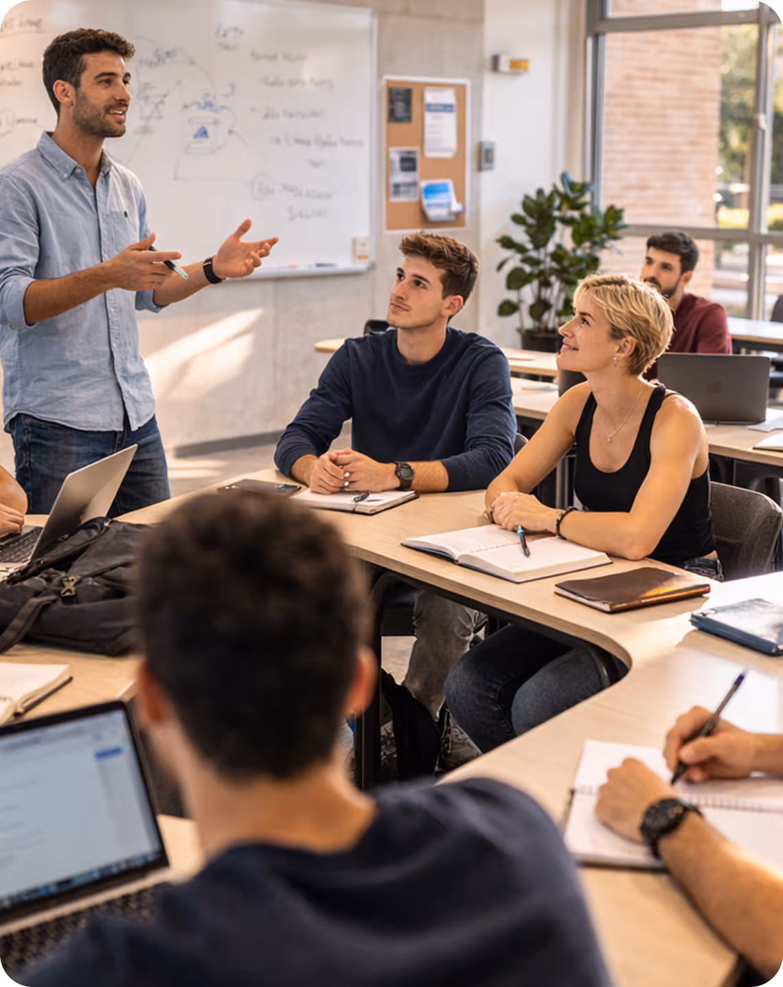 A man standing and speaking to attentive young adults seated around a table in a bright classroom with notebooks and laptops.