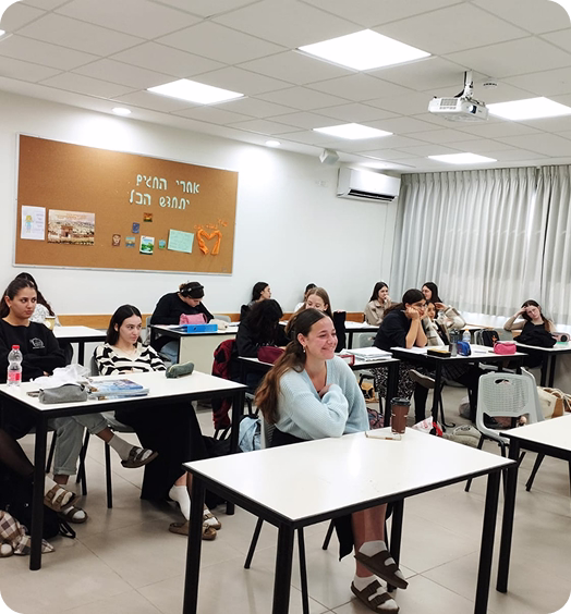 A classroom with adult students sitting at desks, listening attentively, under white fluorescent lights and a projector on the ceiling.