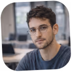 Young man with short curly hair, glasses, and a slight beard, wearing a blue shirt, sitting indoors with blurred background.