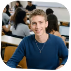 Smiling young man wearing a blue sweater and necklace sitting in a classroom with students in the background.
