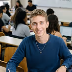 Smiling young man with curly hair wearing a blue sweater sitting at a desk in a classroom.
