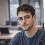 Portrait of a young man with short curly hair and glasses wearing a dark blue shirt in an indoor setting.