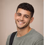 Smiling young man with short curly hair wearing a green shirt and shoulder bag against a plain background.