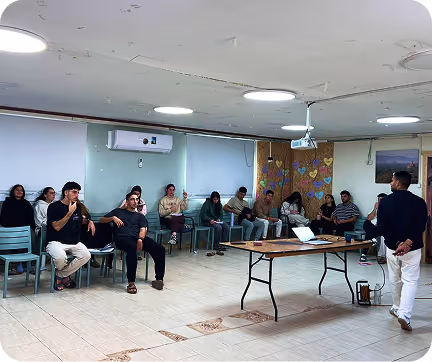 Group of people seated in a classroom setting while a man stands and speaks near a table with a laptop and projector.