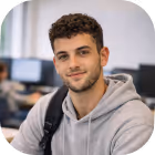 Young man with curly hair wearing a gray hoodie and black backpack, smiling in an office setting.