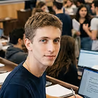 Young man with short brown hair sitting inside a classroom with laptops and students in the background.